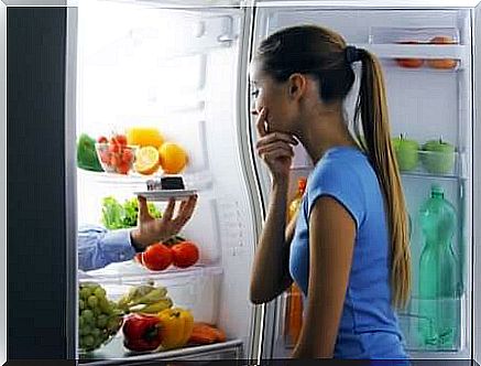 A woman in front of an open fridge hesitating between a piece of fruit and a piece of chocolate cake. 